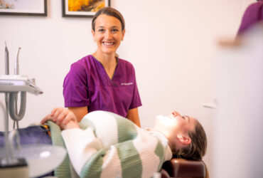 Gratis Zahnspange Vertragsärzte, Dr Kittsteiner Kieferorthopädin lächelt in die Kamera, davor liegt eine Patientin auf einem Stuhl